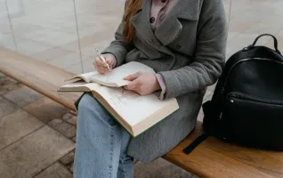 A woman in a gray coat sitting on a bench writing in a notebook outdoors.