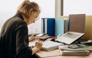Woman in glasses intensely working at a desk with documents and a laptop.