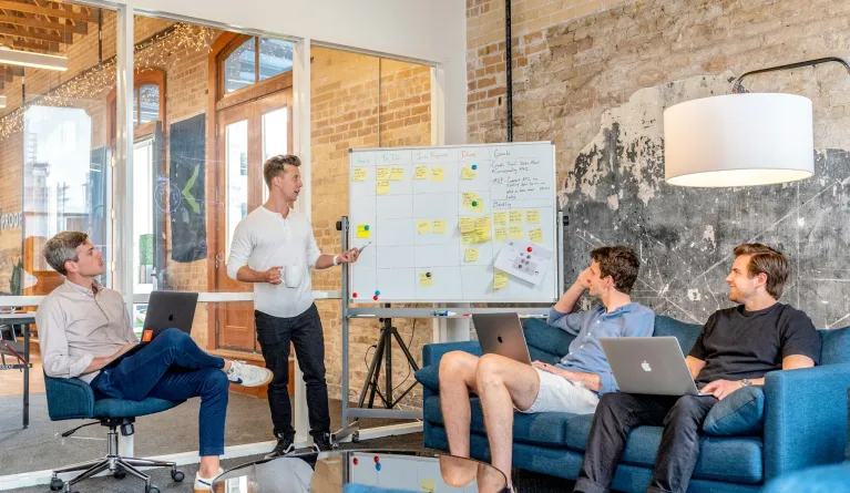 Three men sitting while using laptops and watching man beside whiteboard give employee training