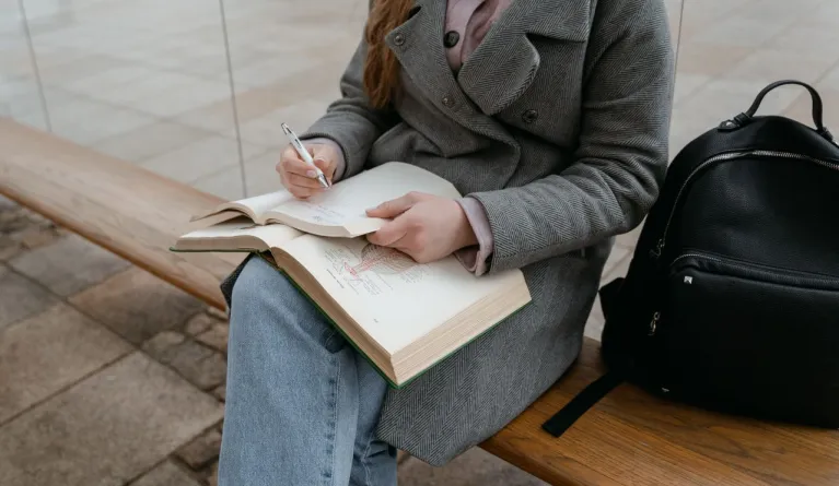 A woman in a gray coat sitting on a bench writing in a notebook outdoors.