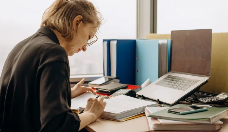 Woman in glasses intensely working at a desk with documents and a laptop.